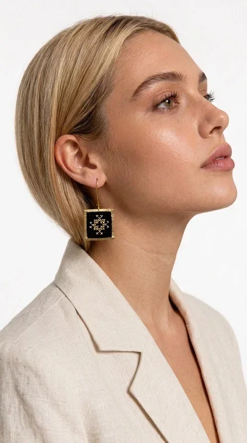 , Embroidered statement earrings with geometric patterns in black are the ideal choice for those seeking jewelry that combines the traditional art of embroidery with a modern, striking result. Designed by Greek Jewelry Designer Aikaterini Chalkiadaki, these handmade earrings are a unique piece of folklore aesthetic that immediately stands out.

Made of brass and fabric, the earrings feature a square, diamond-shaped pendant in deep black, decorated with a golden geometric motif reminiscent of the Greek folk embroidery tradition. Their golden brass metal frame adds elegance and contrast, while the overall result is dynamic yet refined.

These striking dangle earrings suit both casual and formal looks — from an everyday combination with a linen blazer to an evening look for a wedding or christening. They particularly enhance updos and loose hair.