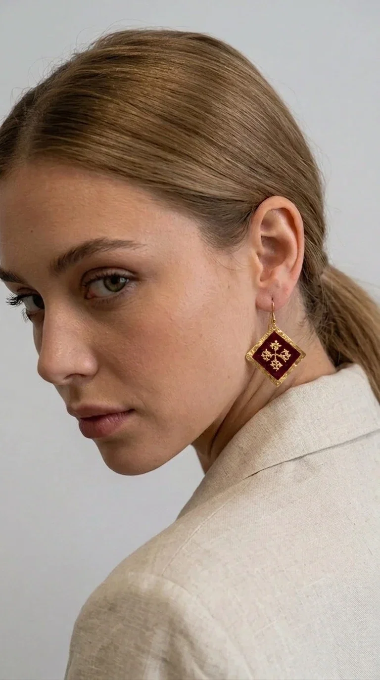 , Embroidered statement earrings with geometric patterns in deep burgundy are the ideal choice for every woman who wants to express herself through her style — with character, color, and a distinctive handmade aesthetic.

Designed by Greek Jewelry Designer Aikaterini Chalkiadaki, these striking dangle earrings combine brass and fabric in a rhombus shape reminiscent of folklore aesthetics and Byzantine tradition. The embroidered floral motif at the center — in golden hues against a deep burgundy background — gives the piece a vintage, boho-chic dimension that suits wedding events, evening outings, and everyday fashion looks alike.

The brass frame with its finely worked gold-toned surface highlights the embroidered fabric, creating a jewel that seems to come from another era — without losing its modern appeal. Lightweight on the ear, hanging from a hook, they pair well with both casual and formal styles, from a linen jacket to an evening dress.