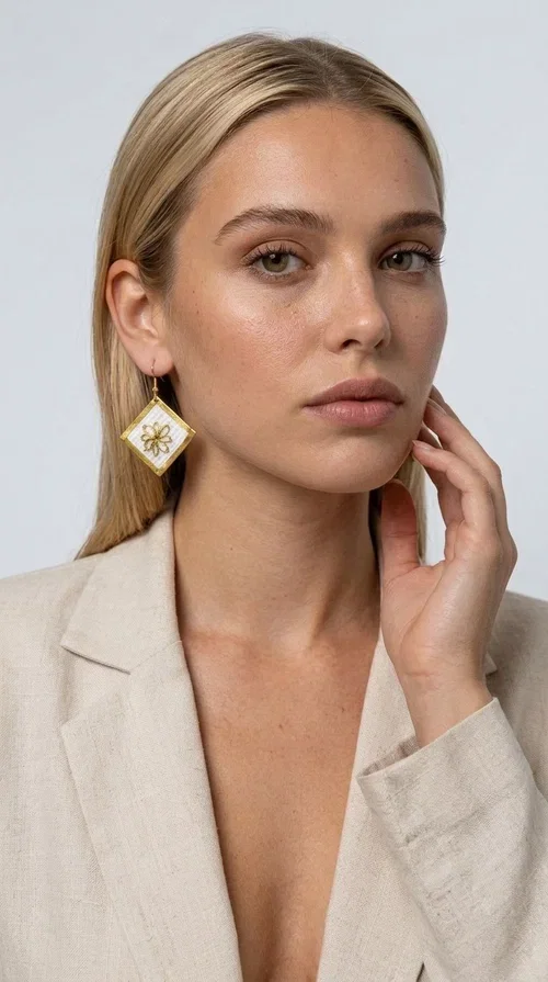 , The embroidered statement earrings with geometric patterns in white and gold are the perfect choice for those seeking a piece of jewelry that combines folk art with contemporary design. Designed by Greek Jewelry Designer Aikaterini Chalkiadaki, these handmade dangle earrings impress with their geometric diamond shape and the embroidered floral decoration at their center.

Made of brass and fabric, they feature a gold frame surrounding a white embroidered section with a raised floral motif — a detail that evokes traditional Greek embroidery art. They hang from a gold hoop, creating an elegant yet striking result. Ideal as earrings for a wedding, christening or any formal event, but also as a boho statement piece for casual looks.