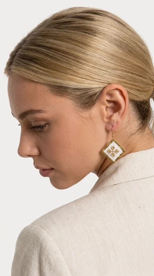 , The embroidered statement earrings with geometric patterns in white and gold are the ideal choice for those seeking a piece of jewelry that combines traditional artistry with contemporary style. Designed by Greek Jewelry Designer Aikaterini Chalkiadaki, these handmade earrings represent a distinctive expression of Greek creativity and craftsmanship.

Their diamond shape, adorned with delicate embroidered decoration on white fabric and gold detailing, evokes the rich tradition of Greek folk art. The combination of brass and fabric creates a piece of jewelry that is light to wear yet striking in presence — perfect for elevating any look, from casual outfits to formal occasions such as weddings or christenings.

These dangling earrings pair perfectly with linen and minimal clothing, adding a boho-ethnic touch of elegance. They also make an excellent gift choice for women who appreciate unique, handmade jewelry with a Greek identity.