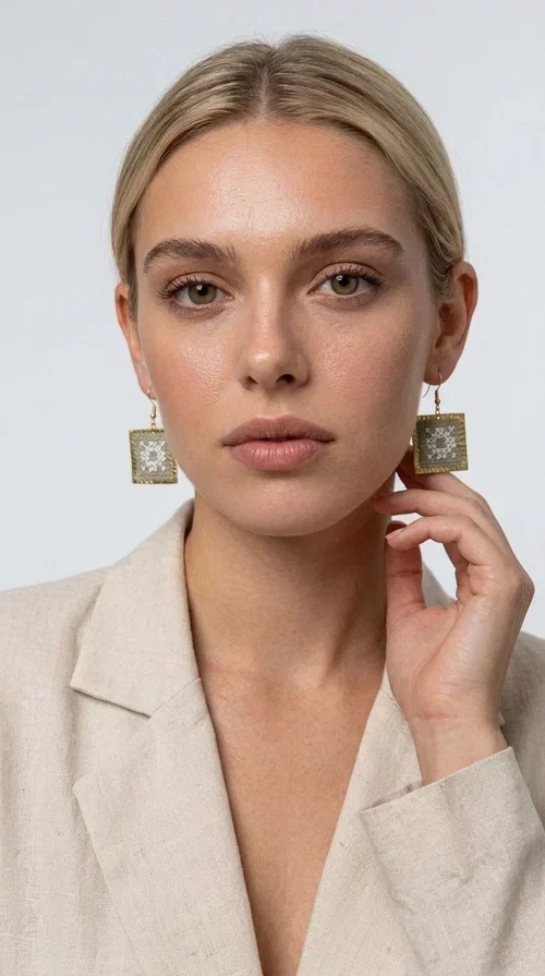 , Embroidered statement earrings with geometric patterns in a cream shade are the ideal choice for anyone seeking an accessory that combines the traditional art of embroidery with a modern, eye-catching aesthetic.

Designed by Greek Jewelry Designer Aikaterini Chalkiadaki, these handmade earrings are a unique combination of brass and fabric. Their square shape in a golden hue elegantly frames the embroidered geometric motif at the center, inspired by Greek folk art and tradition. The warm cream tone of the embroidery exudes a timeless elegance that suits both casual and formal looks equally well.

These striking dangle earrings instantly elevate any outfit — from a simple linen jacket to an evening dress for a wedding or christening. They make a perfect gift for women who appreciate craftsmanship and distinctive design.
