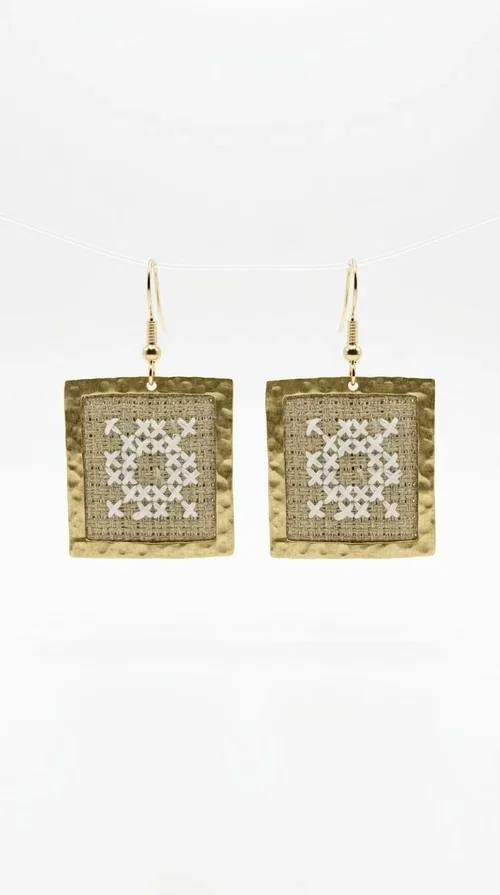 , Embroidered statement earrings with geometric patterns in a cream shade are the ideal choice for anyone seeking an accessory that combines the traditional art of embroidery with a modern, eye-catching aesthetic.

Designed by Greek Jewelry Designer Aikaterini Chalkiadaki, these handmade earrings are a unique combination of brass and fabric. Their square shape in a golden hue elegantly frames the embroidered geometric motif at the center, inspired by Greek folk art and tradition. The warm cream tone of the embroidery exudes a timeless elegance that suits both casual and formal looks equally well.

These striking dangle earrings instantly elevate any outfit — from a simple linen jacket to an evening dress for a wedding or christening. They make a perfect gift for women who appreciate craftsmanship and distinctive design.