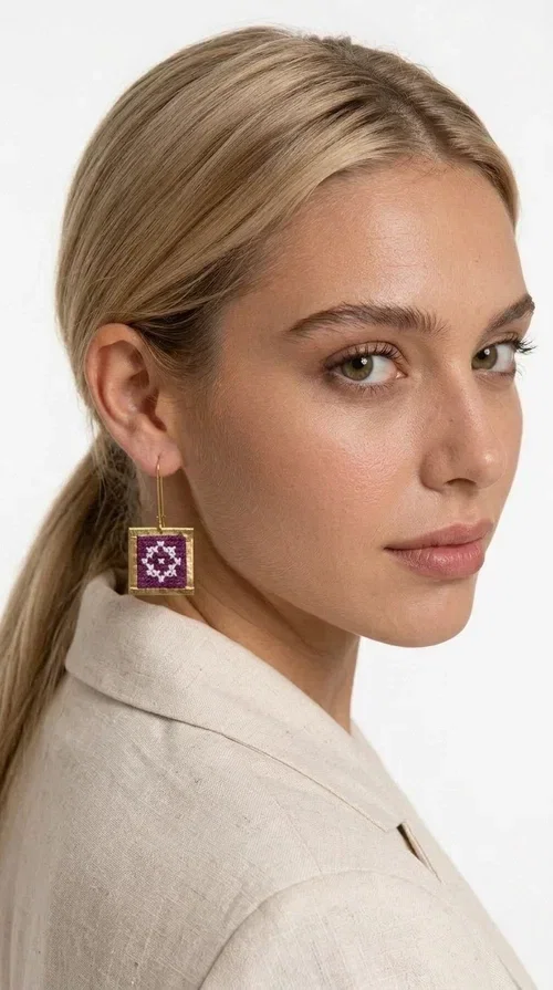 , These embroidered statement earrings with geometric designs in magenta are the ideal choice for those seeking a piece of jewelry that combines folk tradition with contemporary style. Designed by Greek Jewelry Designer Aikaterini Chalkiadaki, these handmade earrings are a distinctive piece that never goes unnoticed.

Each pair features a square shape with a golden brass frame surrounding a fabric panel with traditional cross-stitch embroidery in deep magenta and white. The geometric motif strongly evokes traditional Greek embroidery — a folklore aesthetic that suits both casual and formal looks equally well. They hang from a delicate gold-tone hook, making them comfortable to wear and lightweight on the ear.

Ideal for weddings, christenings, summer outings, and for adding a pop of color to an everyday look. They pair beautifully with neutral outfits — just as shown in the photo, where the vibrant magenta contrasts with a beige jacket for a striking result.