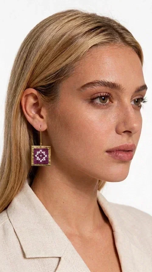 , These embroidered statement earrings with geometric designs in magenta are the ideal choice for those seeking a piece of jewelry that combines folk tradition with contemporary style. Designed by Greek Jewelry Designer Aikaterini Chalkiadaki, these handmade earrings are a distinctive piece that never goes unnoticed.

Each pair features a square shape with a golden brass frame surrounding a fabric panel with traditional cross-stitch embroidery in deep magenta and white. The geometric motif strongly evokes traditional Greek embroidery — a folklore aesthetic that suits both casual and formal looks equally well. They hang from a delicate gold-tone hook, making them comfortable to wear and lightweight on the ear.

Ideal for weddings, christenings, summer outings, and for adding a pop of color to an everyday look. They pair beautifully with neutral outfits — just as shown in the photo, where the vibrant magenta contrasts with a beige jacket for a striking result.