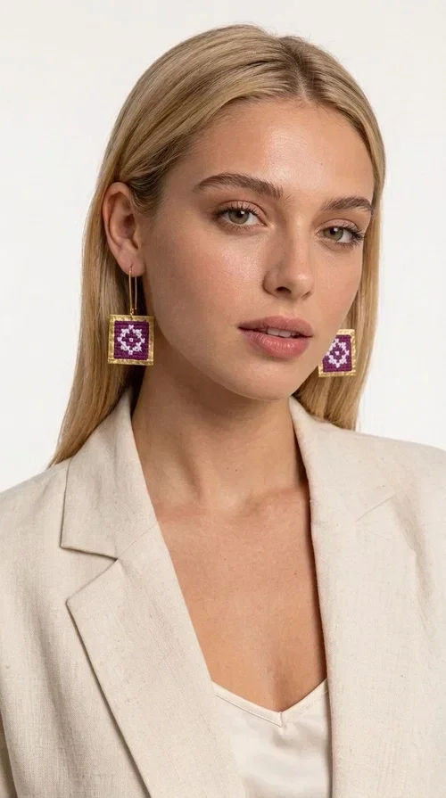 , These embroidered statement earrings with geometric designs in magenta are the ideal choice for those seeking a piece of jewelry that combines folk tradition with contemporary style. Designed by Greek Jewelry Designer Aikaterini Chalkiadaki, these handmade earrings are a distinctive piece that never goes unnoticed.

Each pair features a square shape with a golden brass frame surrounding a fabric panel with traditional cross-stitch embroidery in deep magenta and white. The geometric motif strongly evokes traditional Greek embroidery — a folklore aesthetic that suits both casual and formal looks equally well. They hang from a delicate gold-tone hook, making them comfortable to wear and lightweight on the ear.

Ideal for weddings, christenings, summer outings, and for adding a pop of color to an everyday look. They pair beautifully with neutral outfits — just as shown in the photo, where the vibrant magenta contrasts with a beige jacket for a striking result.