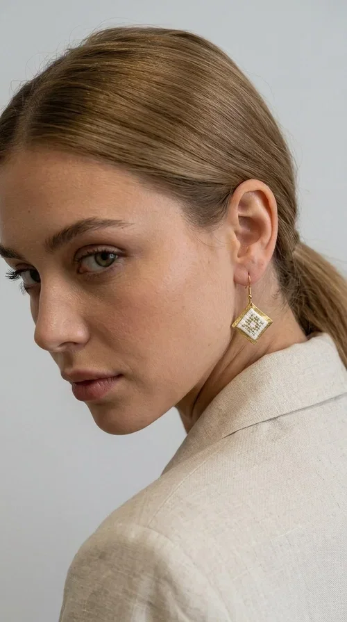 , Embroidered statement earrings with geometric patterns are the ideal choice for every woman who wants to add a folk-chic touch to her look. Designed by Greek Jewelry Designer Aikaterini Chalkiadaki, these handmade earrings combine the tradition of embroidery art with a modern, geometric aesthetic that stands out.

Their diamond shape in white fabric with gold embroidery details and a gold brass frame creates a result that is both elegant and striking. They hang lightly from a gold hook, gently touching the neck and adding movement with every step. Ideal for both everyday wear and special occasions, they pair beautifully with linen and boho outfits, summer dresses, as well as casual-chic looks all year round.