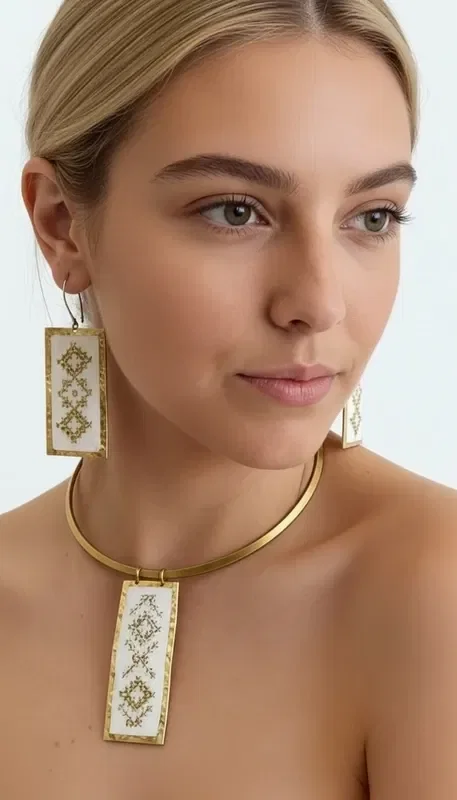 , Discover the art of embroidery in jewelry with this exceptional embroidered white necklace that combines traditional technique with contemporary design. Designed by Greek Jewelry Designer Aikaterini Chalkiadaki, this unique piece features geometric patterns embroidered with gold thread on white fabric, creating a striking contrast that catches the eye.

The statement necklace features a square pendant surface that proudly showcases the handmade geometric motifs, complemented by a gold metal brass collar that adds elegance and stability. The white base with gold thread creates a perfect combination that pairs ideally with summer outfits and bohemian style.

Ideal for both day and evening looks, this embroidered necklace adds an artistic character to your style. It is easy to maintain with gentle cleaning and storage in a dry environment.