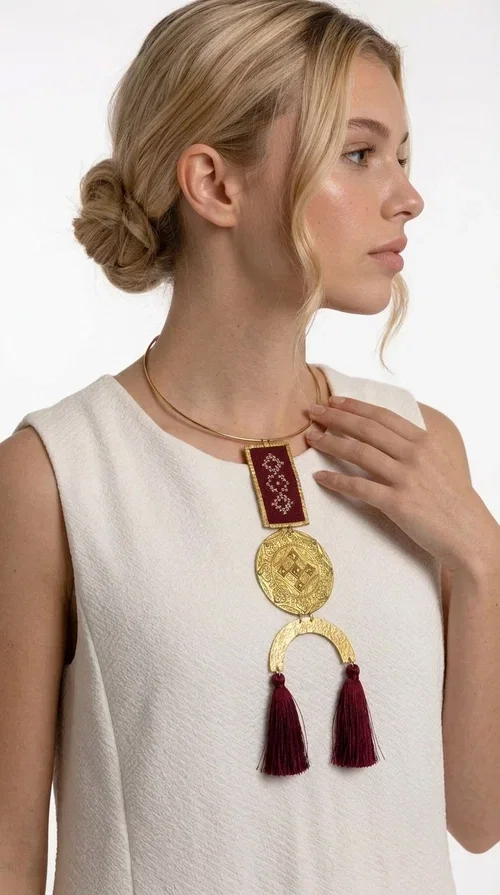 , A statement embroidered necklace that combines tradition with contemporary aesthetics — ideal for every woman who loves striking jewelry with character.

Designed by Greek Jewelry Designer Aikaterini Chalkiadaki, this handmade necklace is a harmonious composition of brass and fabric in a deep burgundy color. The embroidered rectangular piece with folklore-inspired geometric patterns sets the tone, while the round engraved metal piece and the semicircular base with burgundy tassels complete a composition rich in texture and detail. The necklace is worn on a gold-toned torque — clasp-free, lightweight and comfortable to wear.

Ideal for pairing with white linen or a monochrome outfit, it instantly adds character to both casual and evening looks. The geometric embroidery and tassels make it a distinctive piece — a true statement jewelry piece for the neck.