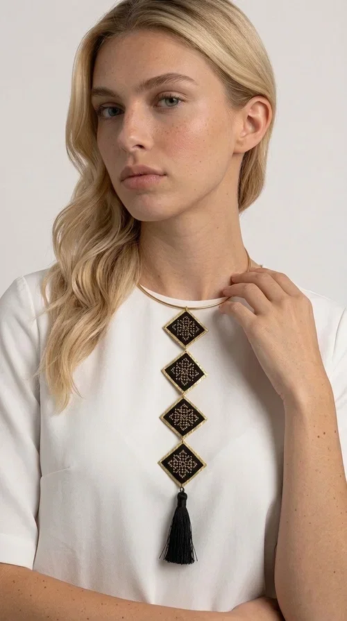 , A statement embroidered necklace that combines tradition with modern aesthetics — ideal for every woman who wants to express herself through her jewelry. Designed by Greek Jewelry Designer Aikaterini Chalkiadaki, this striking long necklace features four diamond-shaped pieces in black fabric with gold-tone geometric embroidered designs, inspired by Greek folk tradition. It is completed with an elegant black tassel at the bottom, while the gold-tone choker base ring adds structure and style.

The necklace is made of brass and fabric, materials that ensure both durability and lightness. The handmade embroidered geometric motifs give each piece a unique character, while the combination of black and gold makes it versatile and timeless. It pairs equally well with casual and formal outfits, instantly offering a bold result.