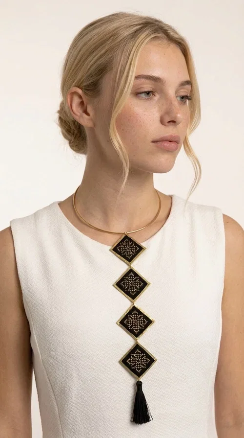 , A statement embroidered necklace that combines tradition with modern aesthetics — ideal for every woman who wants to express herself through her jewelry. Designed by Greek Jewelry Designer Aikaterini Chalkiadaki, this striking long necklace features four diamond-shaped pieces in black fabric with gold-tone geometric embroidered designs, inspired by Greek folk tradition. It is completed with an elegant black tassel at the bottom, while the gold-tone choker base ring adds structure and style.

The necklace is made of brass and fabric, materials that ensure both durability and lightness. The handmade embroidered geometric motifs give each piece a unique character, while the combination of black and gold makes it versatile and timeless. It pairs equally well with casual and formal outfits, instantly offering a bold result.