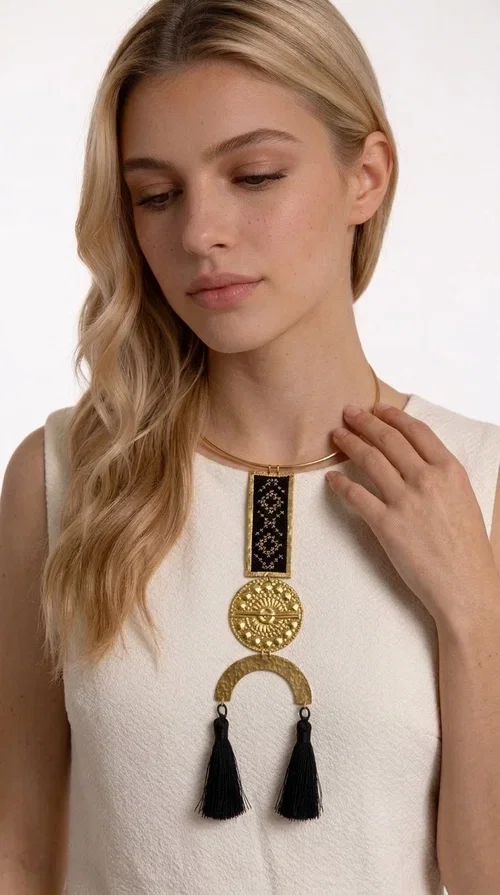 , A statement embroidered necklace that combines tradition with contemporary aesthetics — ideal for those who want to express themselves through their jewelry. Designed by Greek Jewelry Designer Aikaterini Chalkiadaki, this striking necklace is a tribute to folk art and craftsmanship, featuring geometric embroidered designs in deep black that evoke the Greek tradition of embroidery.

The necklace features a rectangular embroidered section with folklore geometric motifs, a gold-tone brass disc with an embossed sun design, a semicircular gold-tone element, and black tassels that add movement and character. It is mounted on a slim gold-tone choker-style chain, creating a silhouette that is both airy and dynamic.

Ideal for summer looks, evening outings, or travel, it pairs beautifully with simple necklines, boho dresses, or linen outfits. Made from brass and fabric, it is lightweight to wear and built to last.
