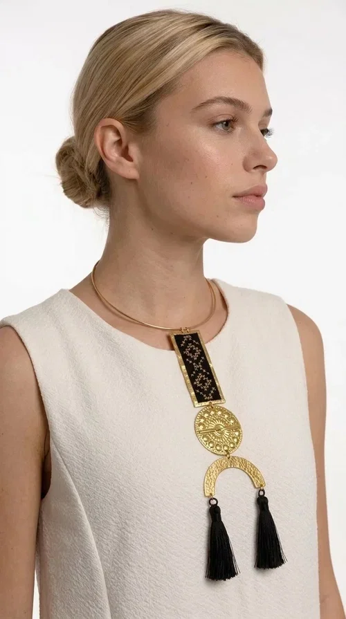 , A statement embroidered necklace that combines tradition with contemporary aesthetics — ideal for those who want to express themselves through their jewelry. Designed by Greek Jewelry Designer Aikaterini Chalkiadaki, this striking necklace is a tribute to folk art and craftsmanship, featuring geometric embroidered designs in deep black that evoke the Greek tradition of embroidery.

The necklace features a rectangular embroidered section with folklore geometric motifs, a gold-tone brass disc with an embossed sun design, a semicircular gold-tone element, and black tassels that add movement and character. It is mounted on a slim gold-tone choker-style chain, creating a silhouette that is both airy and dynamic.

Ideal for summer looks, evening outings, or travel, it pairs beautifully with simple necklines, boho dresses, or linen outfits. Made from brass and fabric, it is lightweight to wear and built to last.