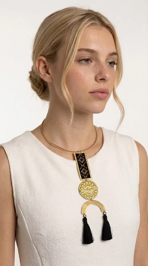 , A statement embroidered necklace that combines tradition with contemporary aesthetics — ideal for those who want to express themselves through their jewelry. Designed by Greek Jewelry Designer Aikaterini Chalkiadaki, this striking necklace is a tribute to folk art and craftsmanship, featuring geometric embroidered designs in deep black that evoke the Greek tradition of embroidery.

The necklace features a rectangular embroidered section with folklore geometric motifs, a gold-tone brass disc with an embossed sun design, a semicircular gold-tone element, and black tassels that add movement and character. It is mounted on a slim gold-tone choker-style chain, creating a silhouette that is both airy and dynamic.

Ideal for summer looks, evening outings, or travel, it pairs beautifully with simple necklines, boho dresses, or linen outfits. Made from brass and fabric, it is lightweight to wear and built to last.