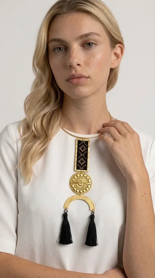 , A statement embroidered necklace that combines tradition with contemporary aesthetics — ideal for those who want to express themselves through their jewelry. Designed by Greek Jewelry Designer Aikaterini Chalkiadaki, this striking necklace is a tribute to folk art and craftsmanship, featuring geometric embroidered designs in deep black that evoke the Greek tradition of embroidery.

The necklace features a rectangular embroidered section with folklore geometric motifs, a gold-tone brass disc with an embossed sun design, a semicircular gold-tone element, and black tassels that add movement and character. It is mounted on a slim gold-tone choker-style chain, creating a silhouette that is both airy and dynamic.

Ideal for summer looks, evening outings, or travel, it pairs beautifully with simple necklines, boho dresses, or linen outfits. Made from brass and fabric, it is lightweight to wear and built to last.
