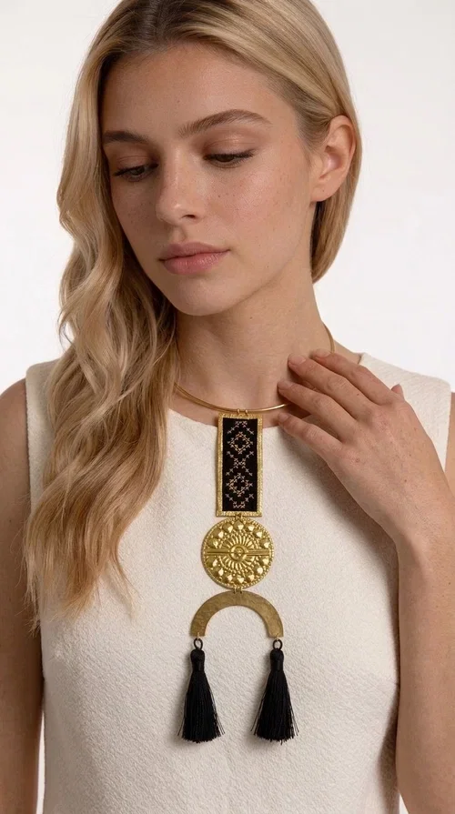 , A statement embroidered necklace that combines tradition with contemporary aesthetics — ideal for those who want to express themselves through their jewelry. Designed by Greek Jewelry Designer Aikaterini Chalkiadaki, this striking necklace is a tribute to folk art and craftsmanship, featuring geometric embroidered designs in deep black that evoke the Greek tradition of embroidery.

The necklace features a rectangular embroidered section with folklore geometric motifs, a gold-tone brass disc with an embossed sun design, a semicircular gold-tone element, and black tassels that add movement and character. It is mounted on a slim gold-tone choker-style chain, creating a silhouette that is both airy and dynamic.

Ideal for summer looks, evening outings, or travel, it pairs beautifully with simple necklines, boho dresses, or linen outfits. Made from brass and fabric, it is lightweight to wear and built to last.