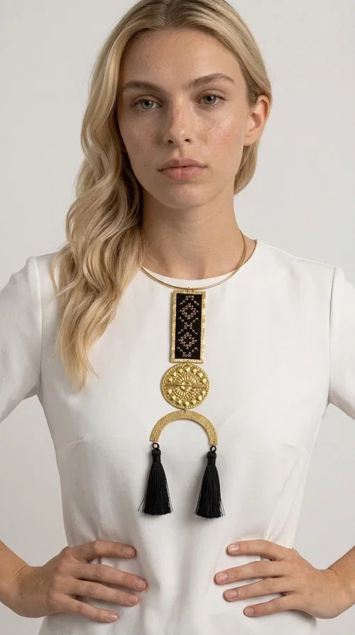 , A statement embroidered necklace that combines tradition with contemporary aesthetics — ideal for those who want to express themselves through their jewelry. Designed by Greek Jewelry Designer Aikaterini Chalkiadaki, this striking necklace is a tribute to folk art and craftsmanship, featuring geometric embroidered designs in deep black that evoke the Greek tradition of embroidery.

The necklace features a rectangular embroidered section with folklore geometric motifs, a gold-tone brass disc with an embossed sun design, a semicircular gold-tone element, and black tassels that add movement and character. It is mounted on a slim gold-tone choker-style chain, creating a silhouette that is both airy and dynamic.

Ideal for summer looks, evening outings, or travel, it pairs beautifully with simple necklines, boho dresses, or linen outfits. Made from brass and fabric, it is lightweight to wear and built to last.