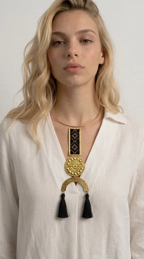 , A statement embroidered necklace that combines tradition with contemporary aesthetics — ideal for those who want to express themselves through their jewelry. Designed by Greek Jewelry Designer Aikaterini Chalkiadaki, this striking necklace is a tribute to folk art and craftsmanship, featuring geometric embroidered designs in deep black that evoke the Greek tradition of embroidery.

The necklace features a rectangular embroidered section with folklore geometric motifs, a gold-tone brass disc with an embossed sun design, a semicircular gold-tone element, and black tassels that add movement and character. It is mounted on a slim gold-tone choker-style chain, creating a silhouette that is both airy and dynamic.

Ideal for summer looks, evening outings, or travel, it pairs beautifully with simple necklines, boho dresses, or linen outfits. Made from brass and fabric, it is lightweight to wear and built to last.