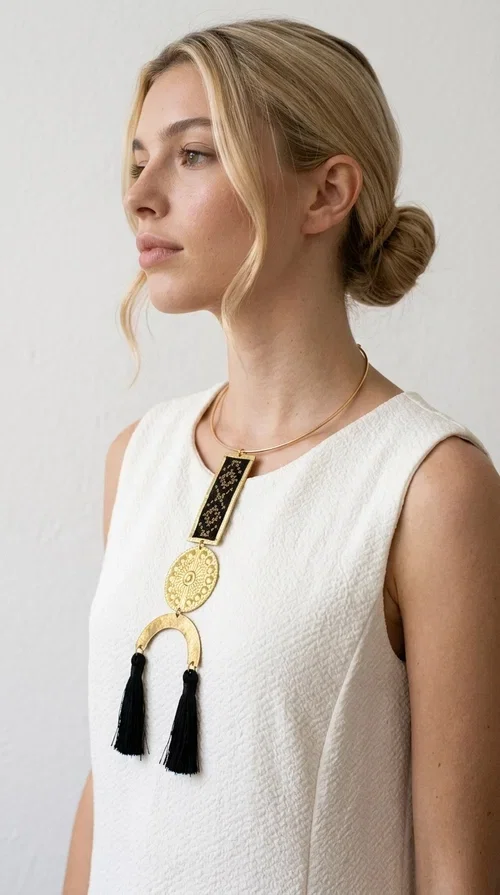 , The statement embroidered necklace with geometric patterns in black is a jewel that never goes unnoticed — bold, handmade, and full of character. Designed by Greek Jewelry Designer Aikaterini Chalkiadaki, it combines the traditional art of embroidery with a modern aesthetic, creating a piece with strong personality and Greek soul.

The design features a gold-tone brass torque collar, from which hangs a rectangular embroidered panel with black geometric motifs inspired by Greek folk tradition. This is followed by a striking embossed brass disc with a sun motif, and at the bottom, a semicircular element adorned with three black tassels, adding movement and femininity to the overall look.

Ideal for looks you want to stand out in — from casual outfits with a linen dress or a plain t-shirt to evening appearances. Black and gold blend harmoniously with almost any clothing color.