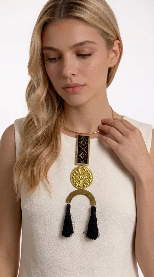 , The statement embroidered necklace with geometric patterns in black is a jewel that never goes unnoticed — bold, handmade, and full of character. Designed by Greek Jewelry Designer Aikaterini Chalkiadaki, it combines the traditional art of embroidery with a modern aesthetic, creating a piece with strong personality and Greek soul.

The design features a gold-tone brass torque collar, from which hangs a rectangular embroidered panel with black geometric motifs inspired by Greek folk tradition. This is followed by a striking embossed brass disc with a sun motif, and at the bottom, a semicircular element adorned with three black tassels, adding movement and femininity to the overall look.

Ideal for looks you want to stand out in — from casual outfits with a linen dress or a plain t-shirt to evening appearances. Black and gold blend harmoniously with almost any clothing color.