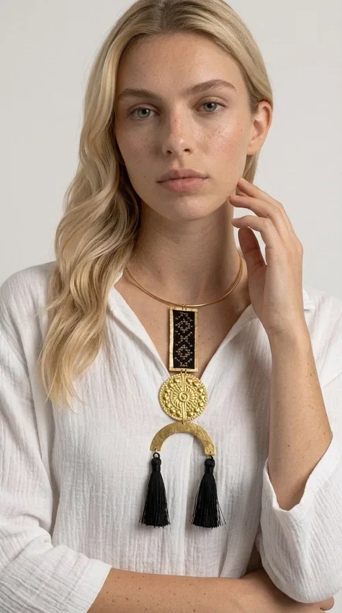 , The statement embroidered necklace with geometric patterns in black is a jewel that never goes unnoticed — bold, handmade, and full of character. Designed by Greek Jewelry Designer Aikaterini Chalkiadaki, it combines the traditional art of embroidery with a modern aesthetic, creating a piece with strong personality and Greek soul.

The design features a gold-tone brass torque collar, from which hangs a rectangular embroidered panel with black geometric motifs inspired by Greek folk tradition. This is followed by a striking embossed brass disc with a sun motif, and at the bottom, a semicircular element adorned with three black tassels, adding movement and femininity to the overall look.

Ideal for looks you want to stand out in — from casual outfits with a linen dress or a plain t-shirt to evening appearances. Black and gold blend harmoniously with almost any clothing color.