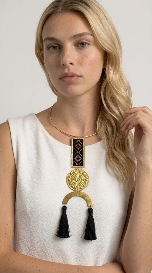 , The statement embroidered necklace with geometric patterns in black is a jewel that never goes unnoticed — bold, handmade, and full of character. Designed by Greek Jewelry Designer Aikaterini Chalkiadaki, it combines the traditional art of embroidery with a modern aesthetic, creating a piece with strong personality and Greek soul.

The design features a gold-tone brass torque collar, from which hangs a rectangular embroidered panel with black geometric motifs inspired by Greek folk tradition. This is followed by a striking embossed brass disc with a sun motif, and at the bottom, a semicircular element adorned with three black tassels, adding movement and femininity to the overall look.

Ideal for looks you want to stand out in — from casual outfits with a linen dress or a plain t-shirt to evening appearances. Black and gold blend harmoniously with almost any clothing color.