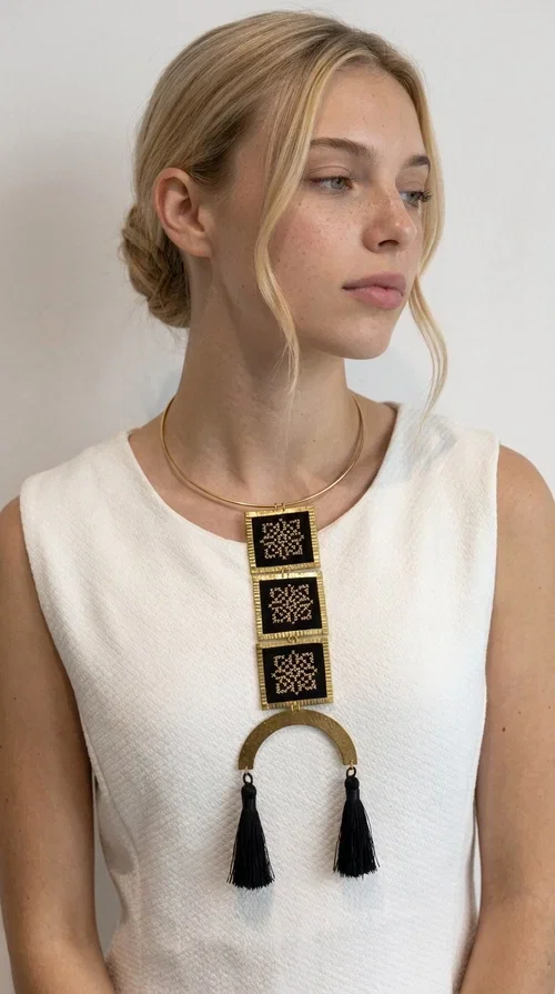 , A statement embroidered necklace that combines the traditional art of embroidery with a modern, striking aesthetic — ideal for those who love jewelry with character and history.

Designed by Greek Jewelry Designer Aikaterini Chalkiadaki, this necklace features three square embroidered elements with geometric patterns in black and gold, inspired by Greek folk tradition. The base of the necklace is a round, snug brass choker in gold tone, from which hangs a series of embroidered squares ending in a semicircular brass element with two striking black tassels. The result is a long, impressive necklace with a strong presence that elevates any outfit.

The geometric patterns of the embroidery evoke a folklore aesthetic, while the combination of fabric and brass gives the piece a unique texture and weight. Ideal for a white or monochrome outfit you want to highlight.