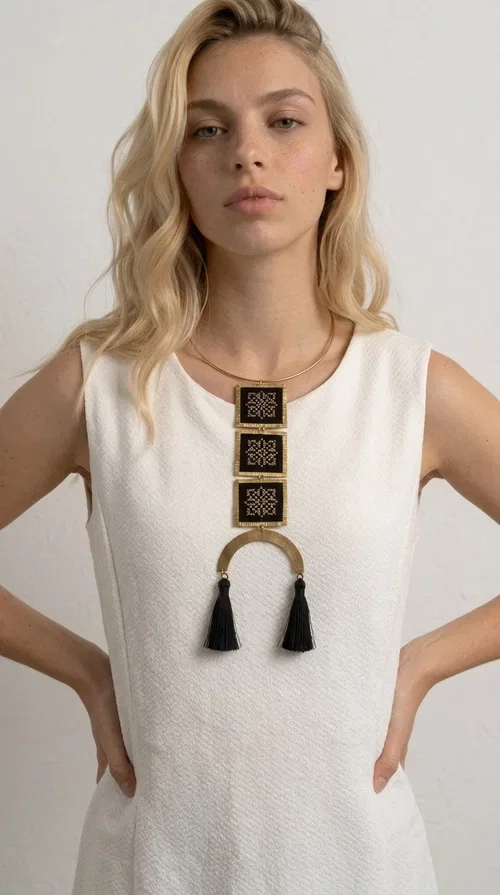 , A statement embroidered necklace that combines the traditional art of embroidery with a modern, striking aesthetic — ideal for those who love jewelry with character and history.

Designed by Greek Jewelry Designer Aikaterini Chalkiadaki, this necklace features three square embroidered elements with geometric patterns in black and gold, inspired by Greek folk tradition. The base of the necklace is a round, snug brass choker in gold tone, from which hangs a series of embroidered squares ending in a semicircular brass element with two striking black tassels. The result is a long, impressive necklace with a strong presence that elevates any outfit.

The geometric patterns of the embroidery evoke a folklore aesthetic, while the combination of fabric and brass gives the piece a unique texture and weight. Ideal for a white or monochrome outfit you want to highlight.