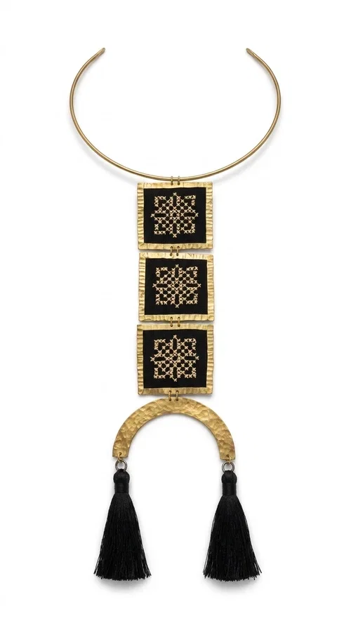 , A statement embroidered necklace that combines the traditional art of embroidery with a modern, striking aesthetic — ideal for those who love jewelry with character and history.

Designed by Greek Jewelry Designer Aikaterini Chalkiadaki, this necklace features three square embroidered elements with geometric patterns in black and gold, inspired by Greek folk tradition. The base of the necklace is a round, snug brass choker in gold tone, from which hangs a series of embroidered squares ending in a semicircular brass element with two striking black tassels. The result is a long, impressive necklace with a strong presence that elevates any outfit.

The geometric patterns of the embroidery evoke a folklore aesthetic, while the combination of fabric and brass gives the piece a unique texture and weight. Ideal for a white or monochrome outfit you want to highlight.