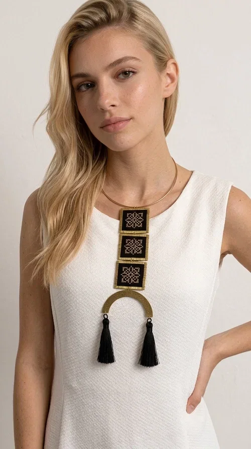 , A statement embroidered necklace that combines the traditional art of embroidery with a modern, striking aesthetic — ideal for those who love jewelry with character and history.

Designed by Greek Jewelry Designer Aikaterini Chalkiadaki, this necklace features three square embroidered elements with geometric patterns in black and gold, inspired by Greek folk tradition. The base of the necklace is a round, snug brass choker in gold tone, from which hangs a series of embroidered squares ending in a semicircular brass element with two striking black tassels. The result is a long, impressive necklace with a strong presence that elevates any outfit.

The geometric patterns of the embroidery evoke a folklore aesthetic, while the combination of fabric and brass gives the piece a unique texture and weight. Ideal for a white or monochrome outfit you want to highlight.