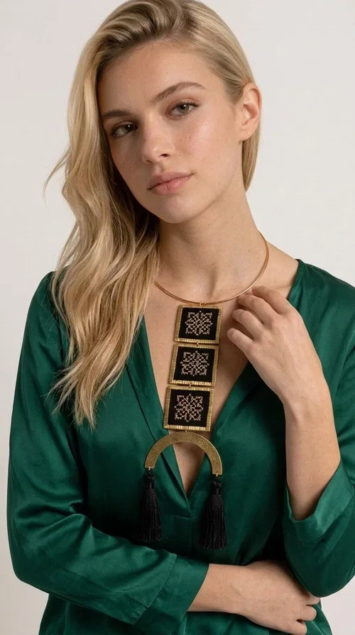, A statement embroidered necklace that combines the traditional art of embroidery with a modern, striking aesthetic — ideal for those who love jewelry with character and history.

Designed by Greek Jewelry Designer Aikaterini Chalkiadaki, this necklace features three square embroidered elements with geometric patterns in black and gold, inspired by Greek folk tradition. The base of the necklace is a round, snug brass choker in gold tone, from which hangs a series of embroidered squares ending in a semicircular brass element with two striking black tassels. The result is a long, impressive necklace with a strong presence that elevates any outfit.

The geometric patterns of the embroidery evoke a folklore aesthetic, while the combination of fabric and brass gives the piece a unique texture and weight. Ideal for a white or monochrome outfit you want to highlight.