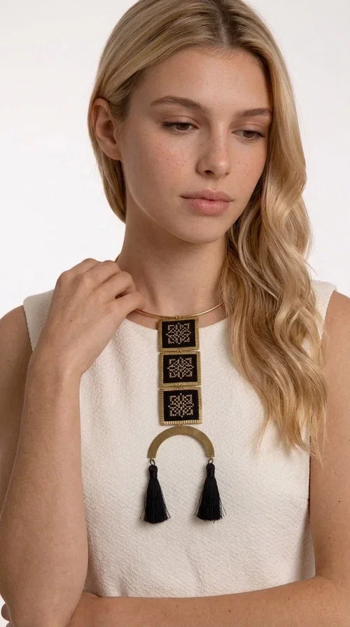 , A statement embroidered necklace that combines the traditional art of embroidery with a modern, striking aesthetic — ideal for those who love jewelry with character and history.

Designed by Greek Jewelry Designer Aikaterini Chalkiadaki, this necklace features three square embroidered elements with geometric patterns in black and gold, inspired by Greek folk tradition. The base of the necklace is a round, snug brass choker in gold tone, from which hangs a series of embroidered squares ending in a semicircular brass element with two striking black tassels. The result is a long, impressive necklace with a strong presence that elevates any outfit.

The geometric patterns of the embroidery evoke a folklore aesthetic, while the combination of fabric and brass gives the piece a unique texture and weight. Ideal for a white or monochrome outfit you want to highlight.