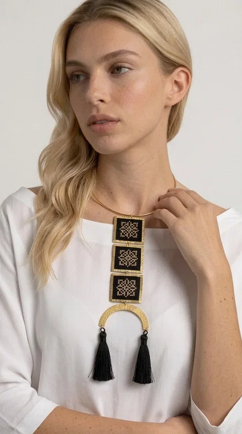 , A statement embroidered necklace that combines the traditional art of embroidery with a modern, striking aesthetic — ideal for those who love jewelry with character and history.

Designed by Greek Jewelry Designer Aikaterini Chalkiadaki, this necklace features three square embroidered elements with geometric patterns in black and gold, inspired by Greek folk tradition. The base of the necklace is a round, snug brass choker in gold tone, from which hangs a series of embroidered squares ending in a semicircular brass element with two striking black tassels. The result is a long, impressive necklace with a strong presence that elevates any outfit.

The geometric patterns of the embroidery evoke a folklore aesthetic, while the combination of fabric and brass gives the piece a unique texture and weight. Ideal for a white or monochrome outfit you want to highlight.