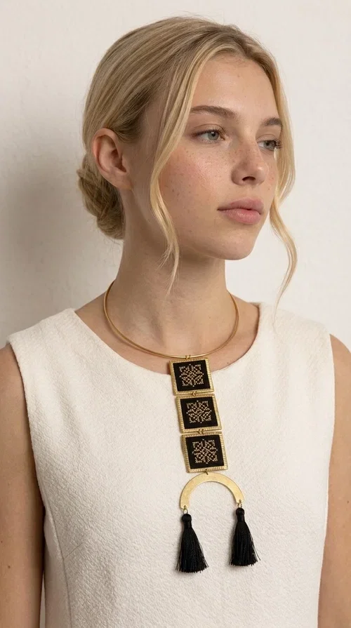 , A statement embroidered necklace that combines the traditional art of embroidery with a modern, striking aesthetic — ideal for those who love jewelry with character and history.

Designed by Greek Jewelry Designer Aikaterini Chalkiadaki, this necklace features three square embroidered elements with geometric patterns in black and gold, inspired by Greek folk tradition. The base of the necklace is a round, snug brass choker in gold tone, from which hangs a series of embroidered squares ending in a semicircular brass element with two striking black tassels. The result is a long, impressive necklace with a strong presence that elevates any outfit.

The geometric patterns of the embroidery evoke a folklore aesthetic, while the combination of fabric and brass gives the piece a unique texture and weight. Ideal for a white or monochrome outfit you want to highlight.