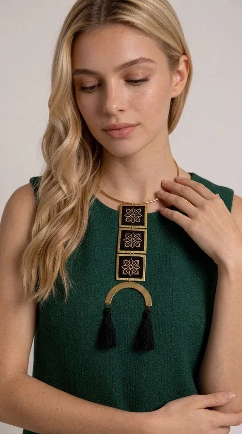 , A statement embroidered necklace that combines the traditional art of embroidery with a modern, striking aesthetic — ideal for those who love jewelry with character and history.

Designed by Greek Jewelry Designer Aikaterini Chalkiadaki, this necklace features three square embroidered elements with geometric patterns in black and gold, inspired by Greek folk tradition. The base of the necklace is a round, snug brass choker in gold tone, from which hangs a series of embroidered squares ending in a semicircular brass element with two striking black tassels. The result is a long, impressive necklace with a strong presence that elevates any outfit.

The geometric patterns of the embroidery evoke a folklore aesthetic, while the combination of fabric and brass gives the piece a unique texture and weight. Ideal for a white or monochrome outfit you want to highlight.