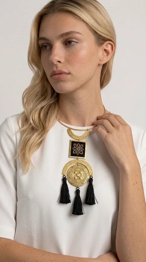 , A statement embroidered necklace that combines tradition with contemporary aesthetics — ideal for those seeking jewelry with character and history.

Designed by Greek Jewelry Designer Aikaterini Chalkiadaki, this striking handmade necklace draws inspiration from folk tradition and the geometric motifs of Greek embroidery. The composition features a hammered semicircular brass frame, an embroidered square section with a black cross-stitch geometric pattern, and an impressive engraved brass disc adorned with traditional relief motifs. At the bottom, four black fabric tassels add movement and depth to the overall design.

The necklace is worn like a torque — a brass hoop that fits comfortably around the neck without a clasp — making it exceptionally easy to wear. It pairs perfectly with minimal or boho outfits, while elevating even a simple monochrome look. Perfect for nights out, celebrations, and for those who want to express their personality in everyday life.