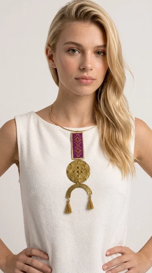 , This statement embroidered necklace combines tradition with modern aesthetics, creating a piece of jewelry that instantly draws attention. Designed by Greek Jewelry Designer Aikaterini Chalkiadaki, it is a handmade creation that exudes a strong folklore character with clever contemporary styling.

The necklace features a rectangular embroidered panel in deep burgundy with geometric patterns, connected to an impressive golden circular brass disc with embossed motifs. The composition is completed with a semicircular element adorned with golden tassels, adding movement and vibrancy. It comes with a slim gold rigid collar that fits comfortably around the neck.

Ideal for elevating a simple white shirt, a boho dress, or even a stylish evening outfit. It instantly adds character and personality to your look.