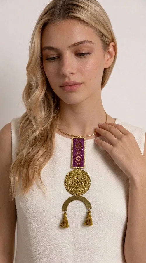 , This statement embroidered necklace combines tradition with modern aesthetics, creating a piece of jewelry that instantly draws attention. Designed by Greek Jewelry Designer Aikaterini Chalkiadaki, it is a handmade creation that exudes a strong folklore character with clever contemporary styling.

The necklace features a rectangular embroidered panel in deep burgundy with geometric patterns, connected to an impressive golden circular brass disc with embossed motifs. The composition is completed with a semicircular element adorned with golden tassels, adding movement and vibrancy. It comes with a slim gold rigid collar that fits comfortably around the neck.

Ideal for elevating a simple white shirt, a boho dress, or even a stylish evening outfit. It instantly adds character and personality to your look.