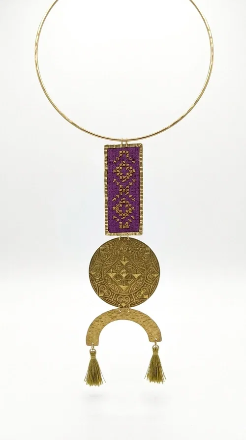 , This statement embroidered necklace combines tradition with modern aesthetics, creating a piece of jewelry that instantly draws attention. Designed by Greek Jewelry Designer Aikaterini Chalkiadaki, it is a handmade creation that exudes a strong folklore character with clever contemporary styling.

The necklace features a rectangular embroidered panel in deep burgundy with geometric patterns, connected to an impressive golden circular brass disc with embossed motifs. The composition is completed with a semicircular element adorned with golden tassels, adding movement and vibrancy. It comes with a slim gold rigid collar that fits comfortably around the neck.

Ideal for elevating a simple white shirt, a boho dress, or even a stylish evening outfit. It instantly adds character and personality to your look.