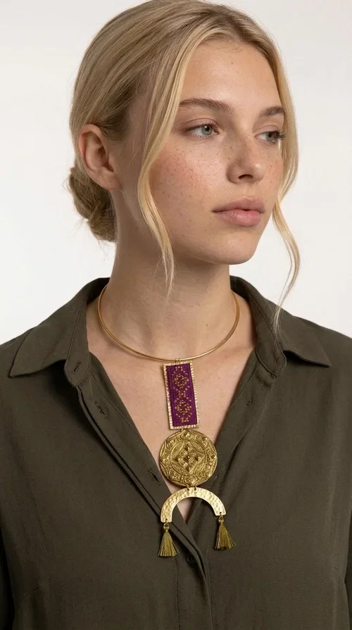 , This statement embroidered necklace combines tradition with modern aesthetics, creating a piece of jewelry that instantly draws attention. Designed by Greek Jewelry Designer Aikaterini Chalkiadaki, it is a handmade creation that exudes a strong folklore character with clever contemporary styling.

The necklace features a rectangular embroidered panel in deep burgundy with geometric patterns, connected to an impressive golden circular brass disc with embossed motifs. The composition is completed with a semicircular element adorned with golden tassels, adding movement and vibrancy. It comes with a slim gold rigid collar that fits comfortably around the neck.

Ideal for elevating a simple white shirt, a boho dress, or even a stylish evening outfit. It instantly adds character and personality to your look.