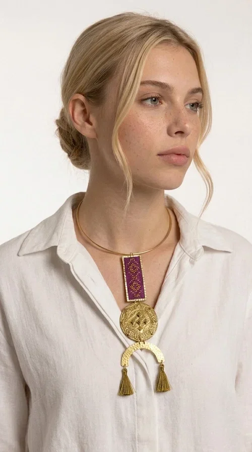 , This statement embroidered necklace combines tradition with modern aesthetics, creating a piece of jewelry that instantly draws attention. Designed by Greek Jewelry Designer Aikaterini Chalkiadaki, it is a handmade creation that exudes a strong folklore character with clever contemporary styling.

The necklace features a rectangular embroidered panel in deep burgundy with geometric patterns, connected to an impressive golden circular brass disc with embossed motifs. The composition is completed with a semicircular element adorned with golden tassels, adding movement and vibrancy. It comes with a slim gold rigid collar that fits comfortably around the neck.

Ideal for elevating a simple white shirt, a boho dress, or even a stylish evening outfit. It instantly adds character and personality to your look.