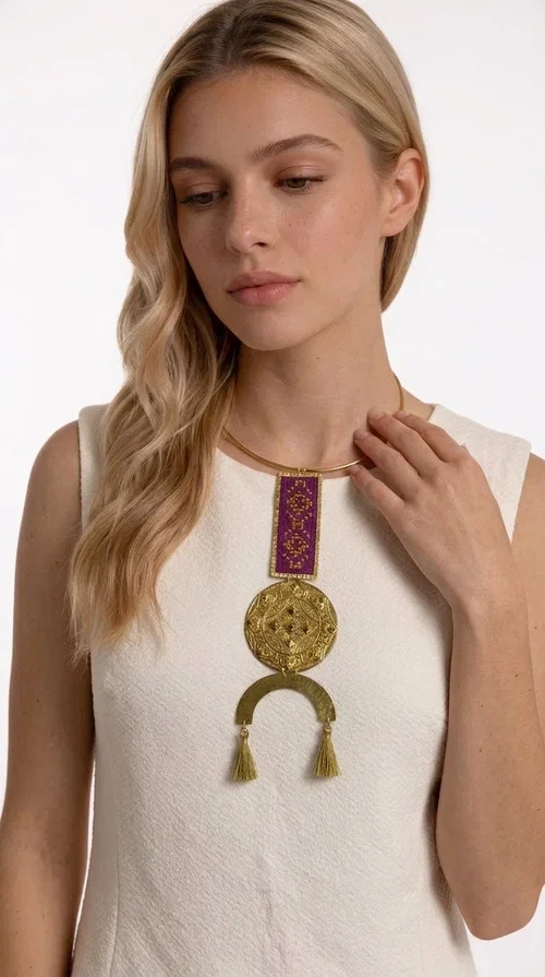 , This statement embroidered necklace combines tradition with modern aesthetics, creating a piece of jewelry that instantly draws attention. Designed by Greek Jewelry Designer Aikaterini Chalkiadaki, it is a handmade creation that exudes a strong folklore character with clever contemporary styling.

The necklace features a rectangular embroidered panel in deep burgundy with geometric patterns, connected to an impressive golden circular brass disc with embossed motifs. The composition is completed with a semicircular element adorned with golden tassels, adding movement and vibrancy. It comes with a slim gold rigid collar that fits comfortably around the neck.

Ideal for elevating a simple white shirt, a boho dress, or even a stylish evening outfit. It instantly adds character and personality to your look.