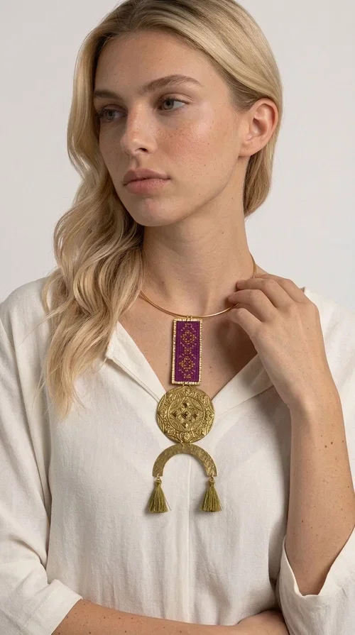 , This statement embroidered necklace combines tradition with modern aesthetics, creating a piece of jewelry that instantly draws attention. Designed by Greek Jewelry Designer Aikaterini Chalkiadaki, it is a handmade creation that exudes a strong folklore character with clever contemporary styling.

The necklace features a rectangular embroidered panel in deep burgundy with geometric patterns, connected to an impressive golden circular brass disc with embossed motifs. The composition is completed with a semicircular element adorned with golden tassels, adding movement and vibrancy. It comes with a slim gold rigid collar that fits comfortably around the neck.

Ideal for elevating a simple white shirt, a boho dress, or even a stylish evening outfit. It instantly adds character and personality to your look.
