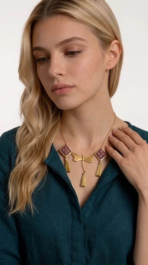 , A statement embroidered necklace that combines the traditional art of embroidery with modern aesthetics — ideal for every woman who wants to express herself with style and originality. Designed by Greek Jewelry Designer Aikaterini Chalkiadaki, this handmade necklace is a true piece of wearable art, inspired by folk heritage and the geometric motifs of Greek tradition.

The necklace features two embroidered square elements in deep burgundy color with gold geometric patterns, joined by a central gold butterfly-shaped motif made of brass. Three gold tassels hang lightly, adding movement and charm to the whole. The gold-tone brass chain completes the elegant, striking look.

It pairs perfectly over simple, solid-colored outfits — such as a blue v-neck top — instantly adding character to your look. Suitable for daytime appearances, nights out, and as a unique gift for women who love striking, feminine jewelry.