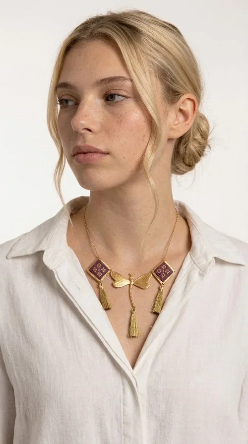 , A statement embroidered necklace that combines the traditional art of embroidery with modern aesthetics — ideal for every woman who wants to express herself with style and originality. Designed by Greek Jewelry Designer Aikaterini Chalkiadaki, this handmade necklace is a true piece of wearable art, inspired by folk heritage and the geometric motifs of Greek tradition.

The necklace features two embroidered square elements in deep burgundy color with gold geometric patterns, joined by a central gold butterfly-shaped motif made of brass. Three gold tassels hang lightly, adding movement and charm to the whole. The gold-tone brass chain completes the elegant, striking look.

It pairs perfectly over simple, solid-colored outfits — such as a blue v-neck top — instantly adding character to your look. Suitable for daytime appearances, nights out, and as a unique gift for women who love striking, feminine jewelry.