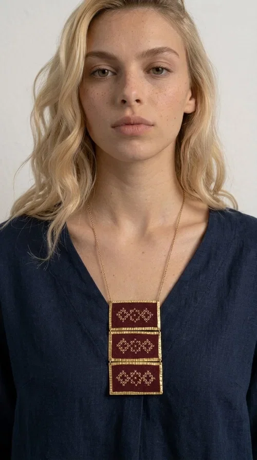 , The statement embroidered necklace in burgundy with geometric patterns is a piece of jewelry that combines the traditional art of embroidery with a modern aesthetic, creating a result that immediately catches the eye. Designed by Greek Jewelry Designer Aikaterini Chalkiadaki, this striking necklace draws inspiration from folk tradition and the geometric motifs that characterize Greek handicraft. The deep burgundy color of the fabric, combined with gold brass, creates a rich, warm palette that suits every skin tone.

The necklace consists of three rectangular embroidered sections, placed one below the other and framed with gold brass featuring a textured finish. Each section showcases handmade embroidery with repeating geometric patterns in a folklore style, reminiscent of traditional Greek embroidery. The result is a long necklace with distinctive character, ideal for elevating a simple outfit or completing a boho-chic look.