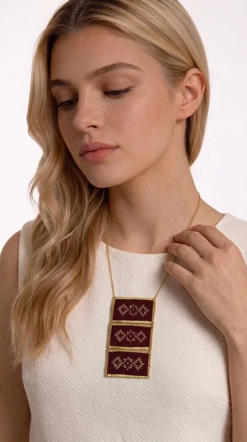 , The statement embroidered necklace in burgundy with geometric patterns is a piece of jewelry that combines the traditional art of embroidery with a modern aesthetic, creating a result that immediately catches the eye. Designed by Greek Jewelry Designer Aikaterini Chalkiadaki, this striking necklace draws inspiration from folk tradition and the geometric motifs that characterize Greek handicraft. The deep burgundy color of the fabric, combined with gold brass, creates a rich, warm palette that suits every skin tone.

The necklace consists of three rectangular embroidered sections, placed one below the other and framed with gold brass featuring a textured finish. Each section showcases handmade embroidery with repeating geometric patterns in a folklore style, reminiscent of traditional Greek embroidery. The result is a long necklace with distinctive character, ideal for elevating a simple outfit or completing a boho-chic look.