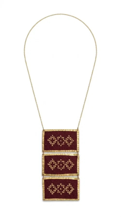 , The statement embroidered necklace in burgundy with geometric patterns is a piece of jewelry that combines the traditional art of embroidery with a modern aesthetic, creating a result that immediately catches the eye. Designed by Greek Jewelry Designer Aikaterini Chalkiadaki, this striking necklace draws inspiration from folk tradition and the geometric motifs that characterize Greek handicraft. The deep burgundy color of the fabric, combined with gold brass, creates a rich, warm palette that suits every skin tone.

The necklace consists of three rectangular embroidered sections, placed one below the other and framed with gold brass featuring a textured finish. Each section showcases handmade embroidery with repeating geometric patterns in a folklore style, reminiscent of traditional Greek embroidery. The result is a long necklace with distinctive character, ideal for elevating a simple outfit or completing a boho-chic look.