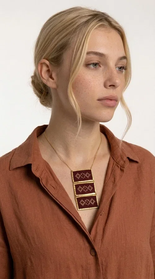, The statement embroidered necklace in burgundy with geometric patterns is a piece of jewelry that combines the traditional art of embroidery with a modern aesthetic, creating a result that immediately catches the eye. Designed by Greek Jewelry Designer Aikaterini Chalkiadaki, this striking necklace draws inspiration from folk tradition and the geometric motifs that characterize Greek handicraft. The deep burgundy color of the fabric, combined with gold brass, creates a rich, warm palette that suits every skin tone.

The necklace consists of three rectangular embroidered sections, placed one below the other and framed with gold brass featuring a textured finish. Each section showcases handmade embroidery with repeating geometric patterns in a folklore style, reminiscent of traditional Greek embroidery. The result is a long necklace with distinctive character, ideal for elevating a simple outfit or completing a boho-chic look.