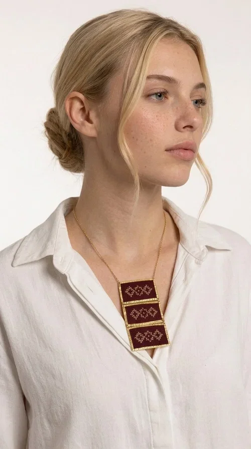 , The statement embroidered necklace in burgundy with geometric patterns is a piece of jewelry that combines the traditional art of embroidery with a modern aesthetic, creating a result that immediately catches the eye. Designed by Greek Jewelry Designer Aikaterini Chalkiadaki, this striking necklace draws inspiration from folk tradition and the geometric motifs that characterize Greek handicraft. The deep burgundy color of the fabric, combined with gold brass, creates a rich, warm palette that suits every skin tone.

The necklace consists of three rectangular embroidered sections, placed one below the other and framed with gold brass featuring a textured finish. Each section showcases handmade embroidery with repeating geometric patterns in a folklore style, reminiscent of traditional Greek embroidery. The result is a long necklace with distinctive character, ideal for elevating a simple outfit or completing a boho-chic look.