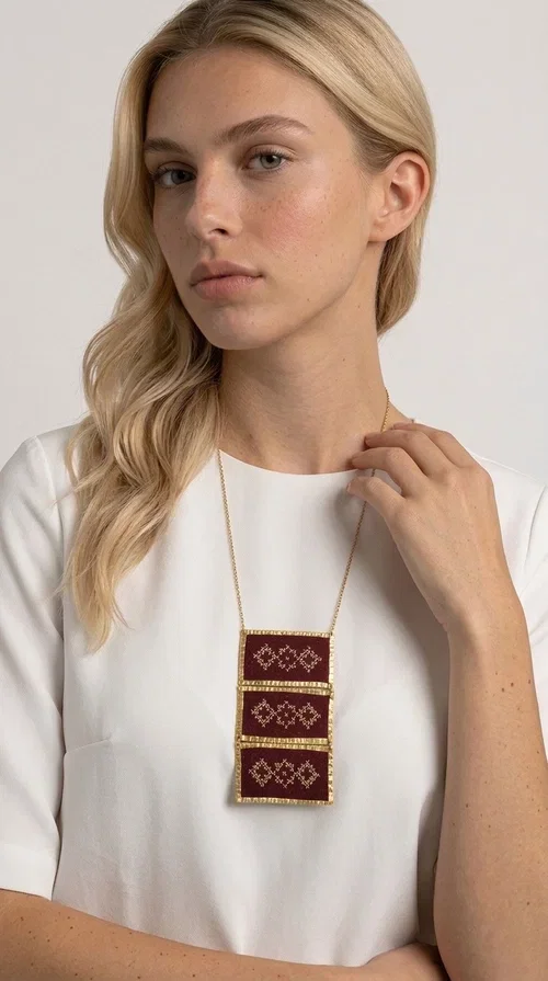 , The statement embroidered necklace in burgundy with geometric patterns is a piece of jewelry that combines the traditional art of embroidery with a modern aesthetic, creating a result that immediately catches the eye. Designed by Greek Jewelry Designer Aikaterini Chalkiadaki, this striking necklace draws inspiration from folk tradition and the geometric motifs that characterize Greek handicraft. The deep burgundy color of the fabric, combined with gold brass, creates a rich, warm palette that suits every skin tone.

The necklace consists of three rectangular embroidered sections, placed one below the other and framed with gold brass featuring a textured finish. Each section showcases handmade embroidery with repeating geometric patterns in a folklore style, reminiscent of traditional Greek embroidery. The result is a long necklace with distinctive character, ideal for elevating a simple outfit or completing a boho-chic look.